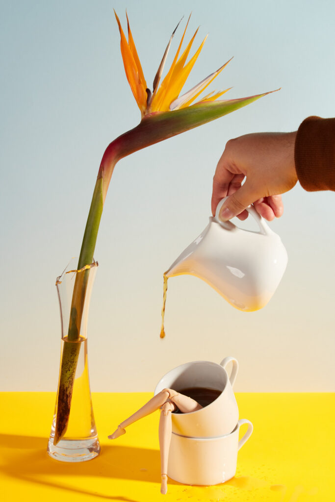 coffee being poured into a cup with doll legs, next to bird of paradise flower. All on a bright yellow table cloth