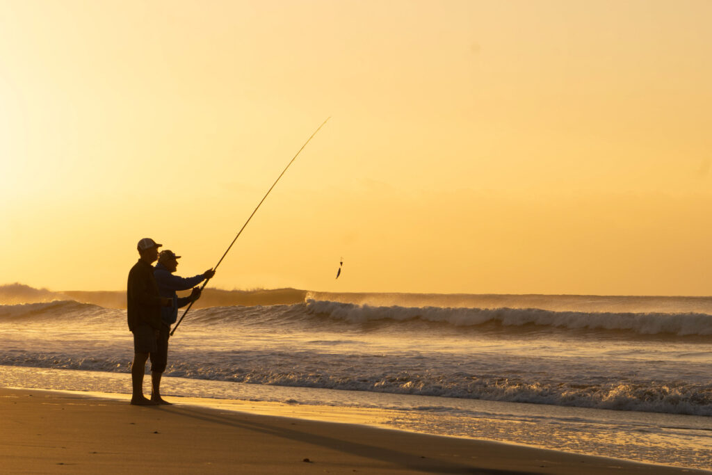 two people fishing on the ocean shore at sunset