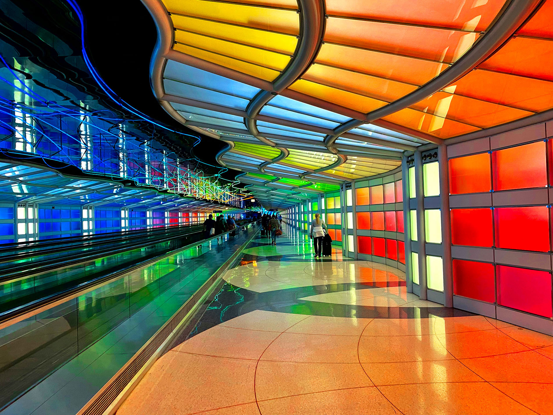 Indoor walkway with moving sidewalk on left. Surrounded by a vibrant rainbow colored panels in walls and ceiling