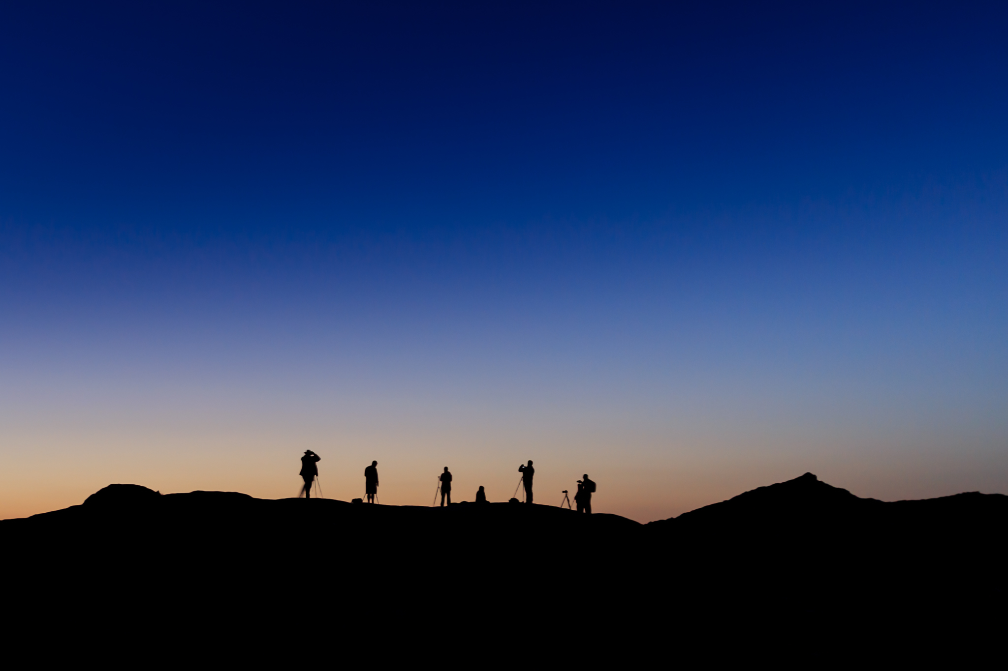 Silhouette of 7 photographers with tripods facing a predawn orange sky that fades to dark blue at Valley of Fire State Park, Nevada